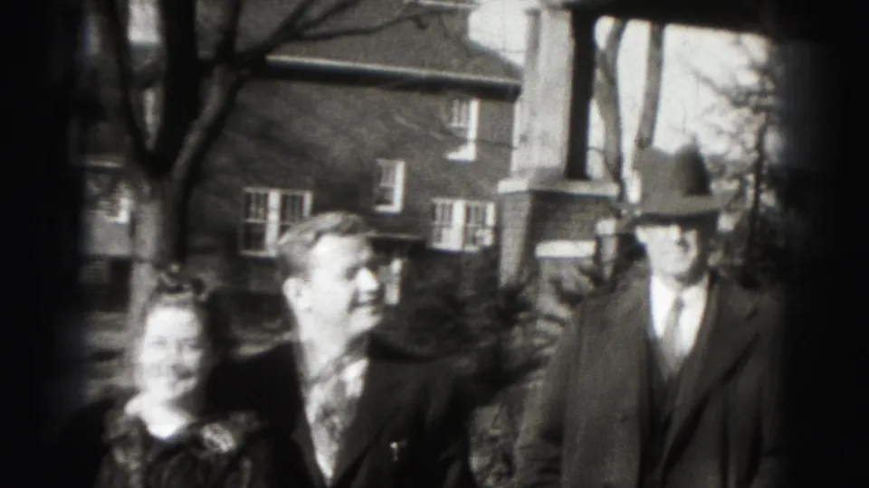 1946 Harrisburg home-movie frame showing three adults in formal clothing outside a brick house with a porch.