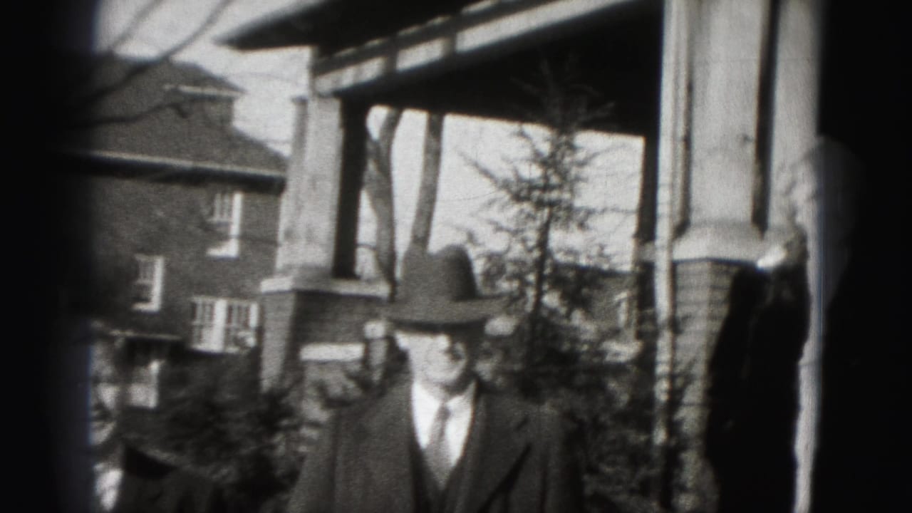 A suited man in a fedora stands outside a Harrisburg brick home, with a second person partly cropped at the left edge.