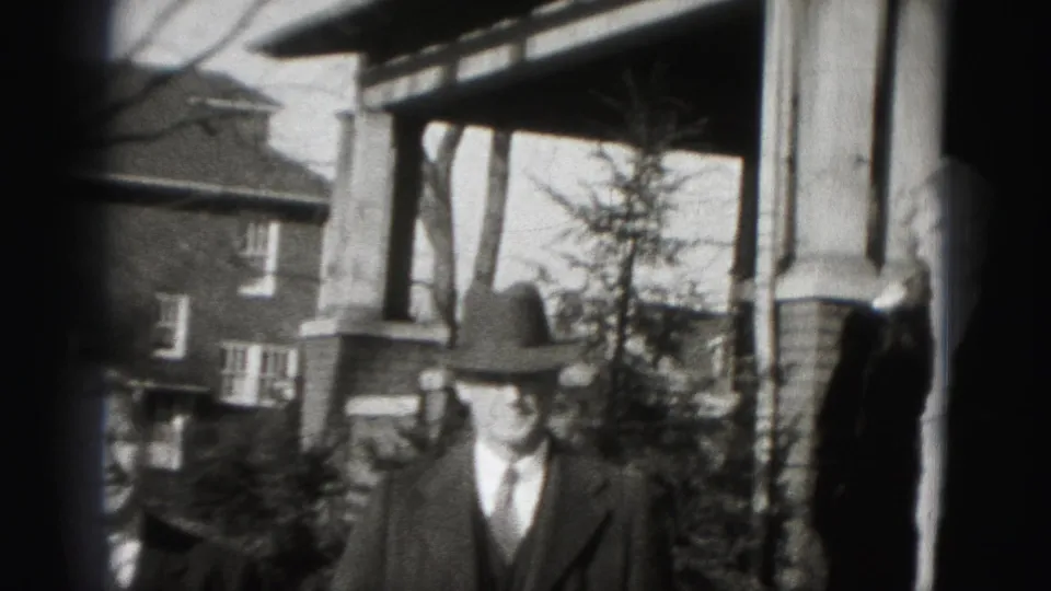 A suited man in a fedora stands outside a Harrisburg brick home, with a second person partly cropped at the left edge.