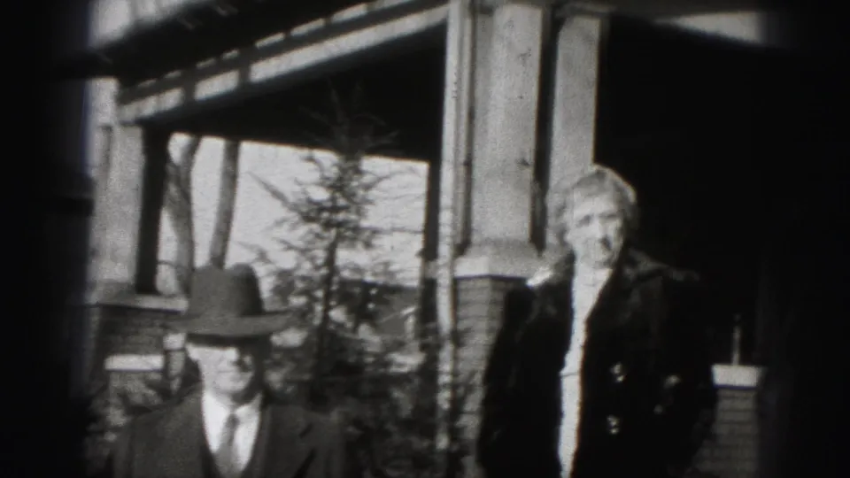 Home-movie frame of a man in a fedora and a woman in a dark coat standing outside a brick house in Harrisburg.
