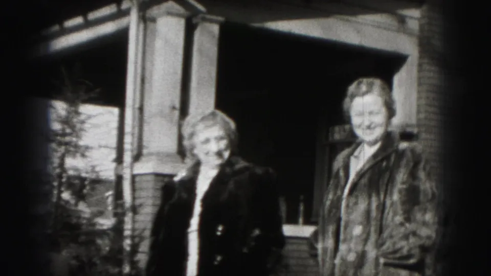 Two women in coats stand outside the Harrisburg brick home, framed by the porch and doorway in a 1946 home movie.