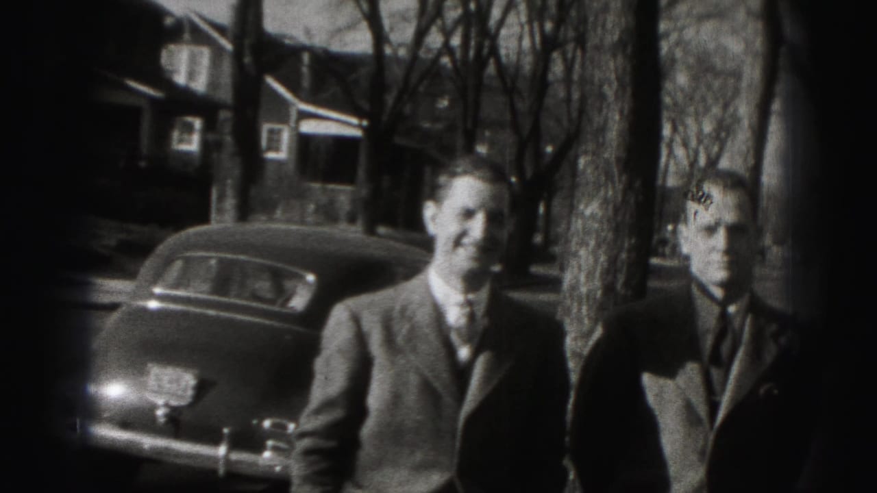 Two suited men stand on a residential street near a parked car and bare trees.