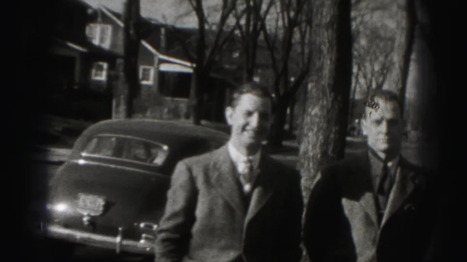 Two men in suits stand on a Harrisburg residential street in 1946 near a parked mid-century car and leafless trees.