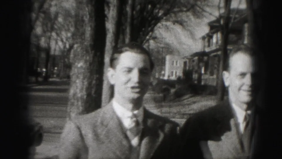 Two suited men smile in a 1946 Harrisburg home-movie frame with bare trees and residential houses behind them.