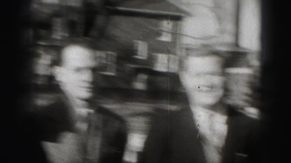 Two suited adult men move through a Harrisburg neighborhood, with brick houses and leafless trees in the background.