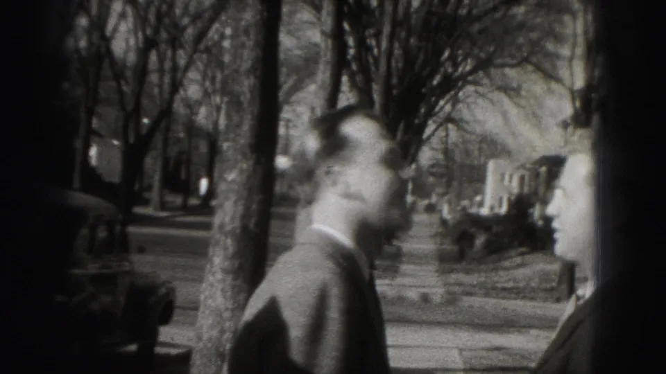 A suited man turns on a Harrisburg sidewalk beside bare trees, parked cars, and another partially visible adult figure.