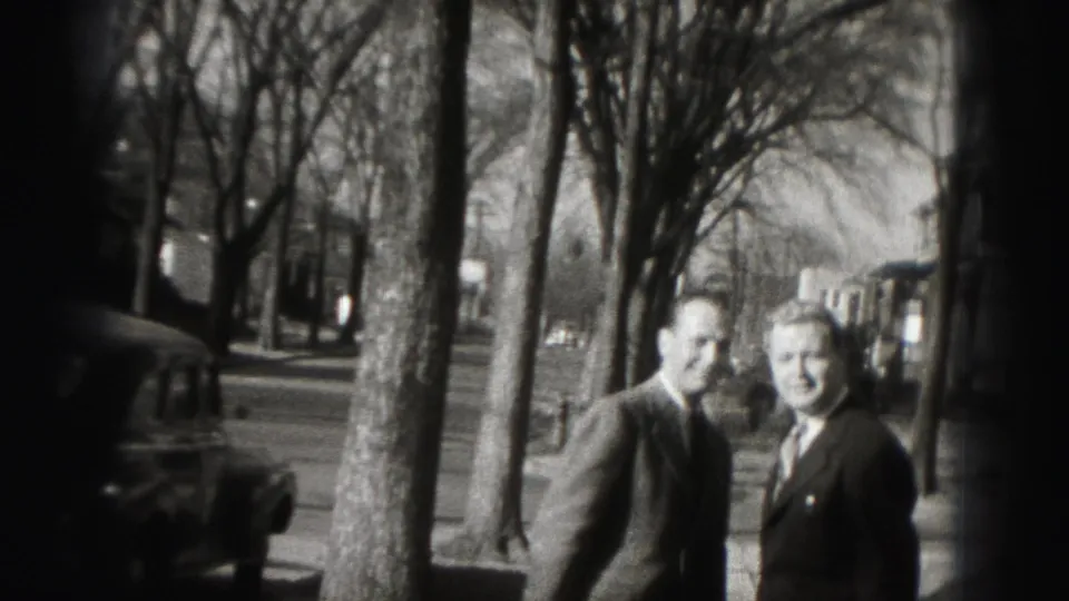 Two suited men turn back toward the camera on a Harrisburg sidewalk, with trees and parked cars behind them.