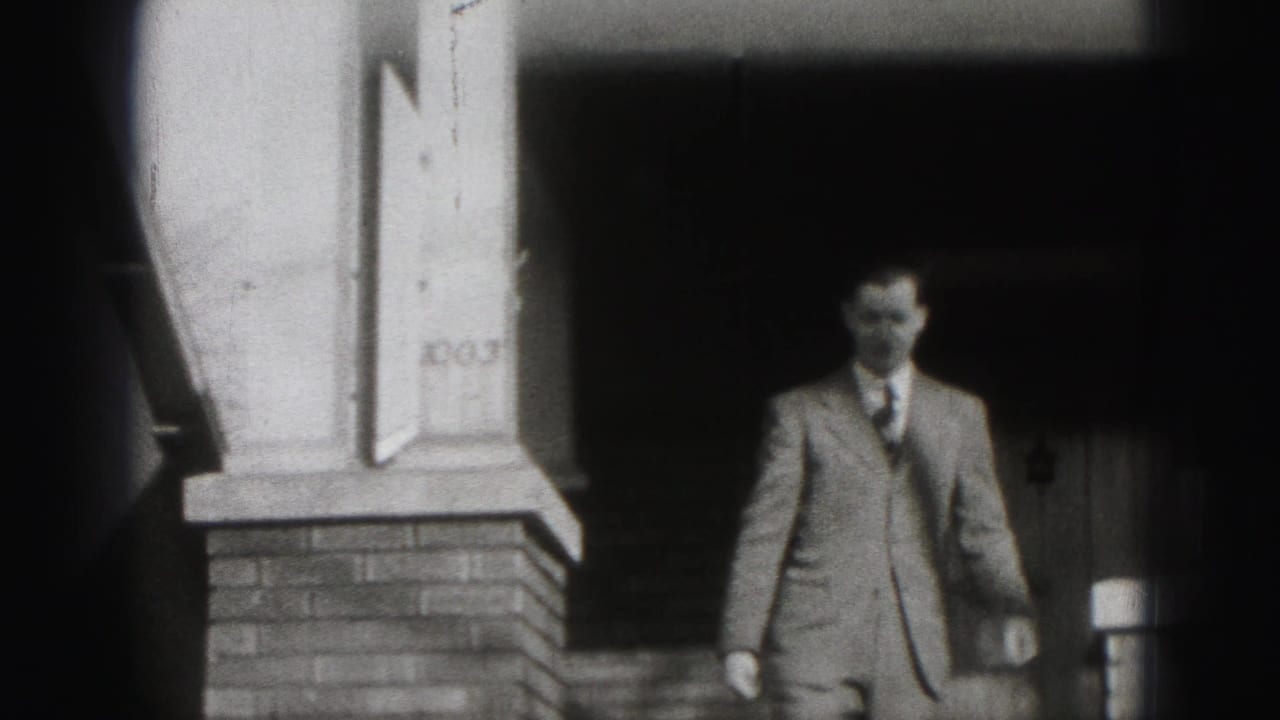 Suited man standing near a brick porch with house number 1003 visible.