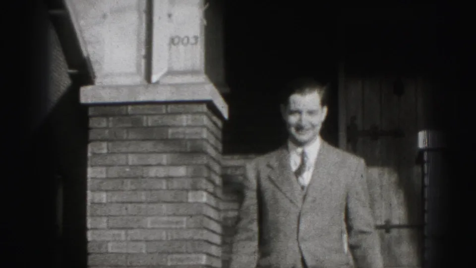 A suited man smiles toward the camera beside a brick residential porch in 1946 Harrisburg home-movie footage.