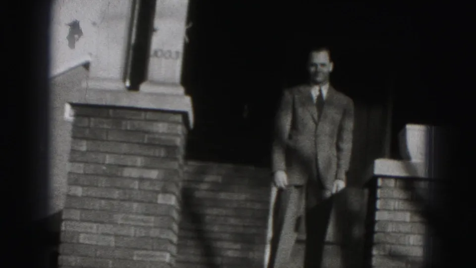 The suited man stands on the porch near the doorway and railing, with the house number 1003 visible above the brick column.