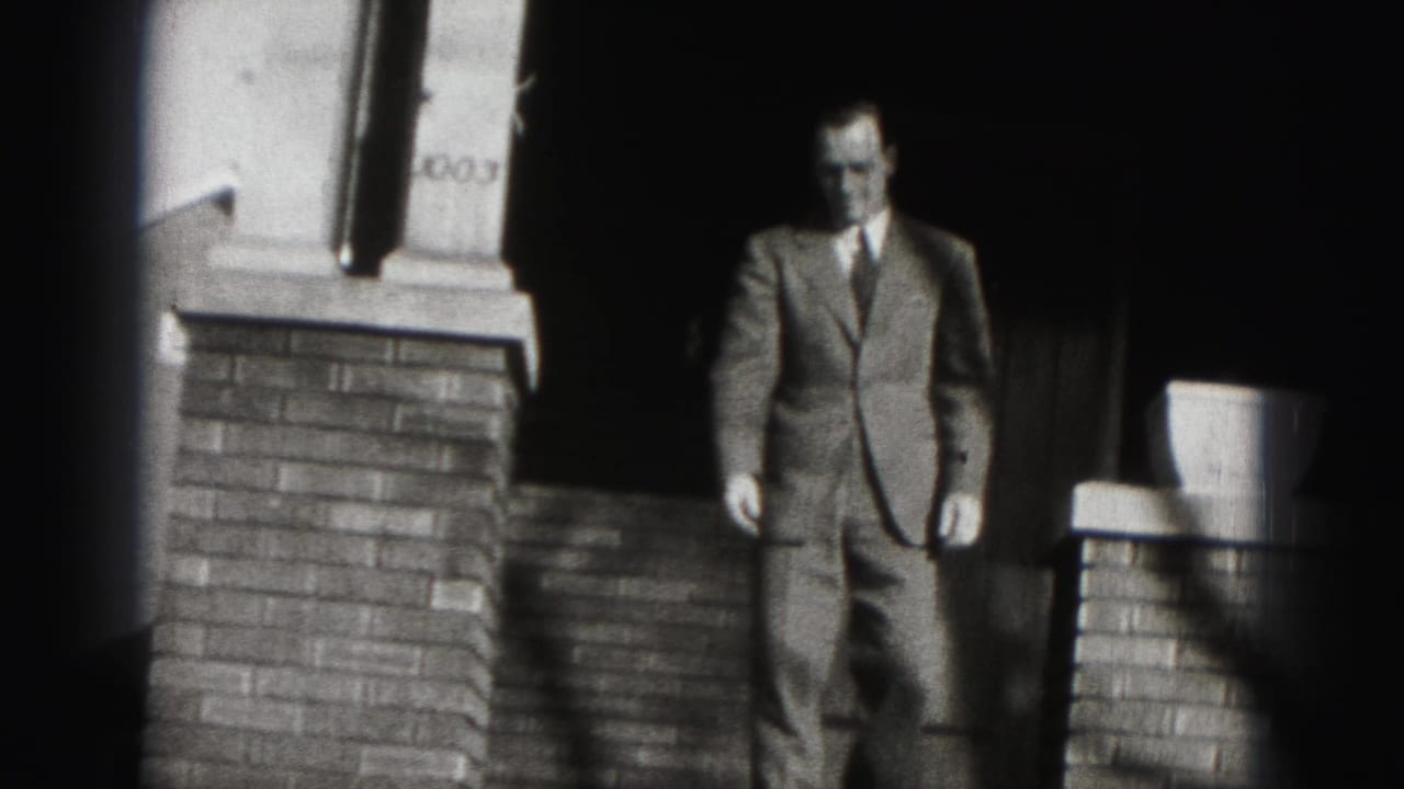 Man in a suit stands in a dark doorway above brick steps.