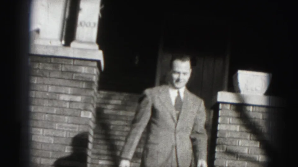 The suited man descends the brick steps of a Harrisburg residence, lit by strong contrast in black-and-white 8mm footage.