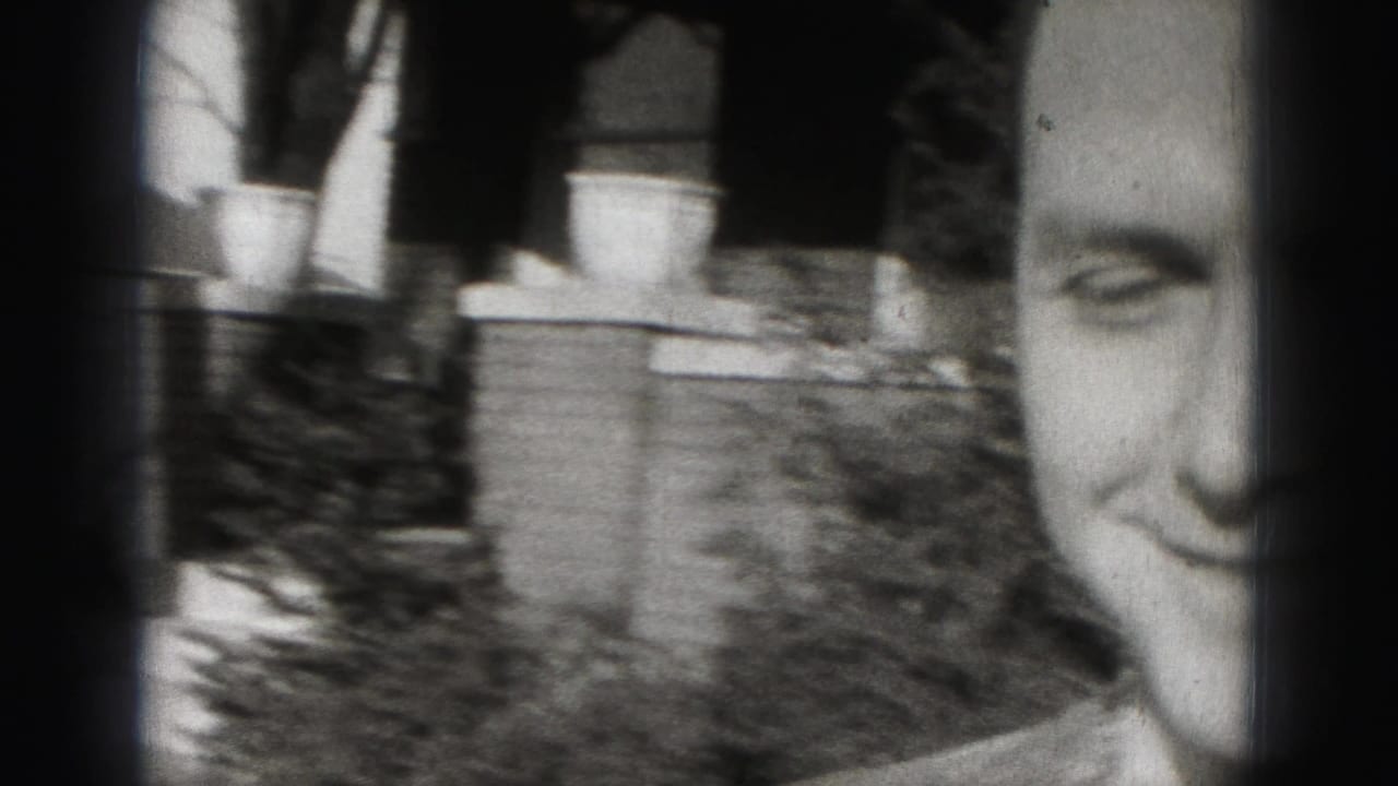 Close view of a smiling adult man beside a porch and shrubs, partly obscured by film-edge shadow.