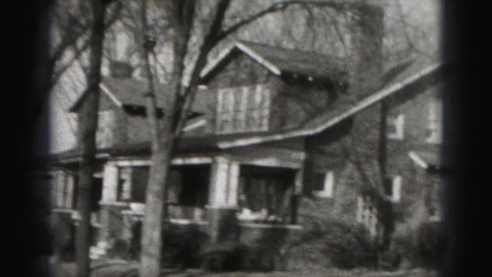 Black-and-white 8mm frame of a Harrisburg residential house with porch, bare trees, and shrubs, framed by dark film-edge shading.