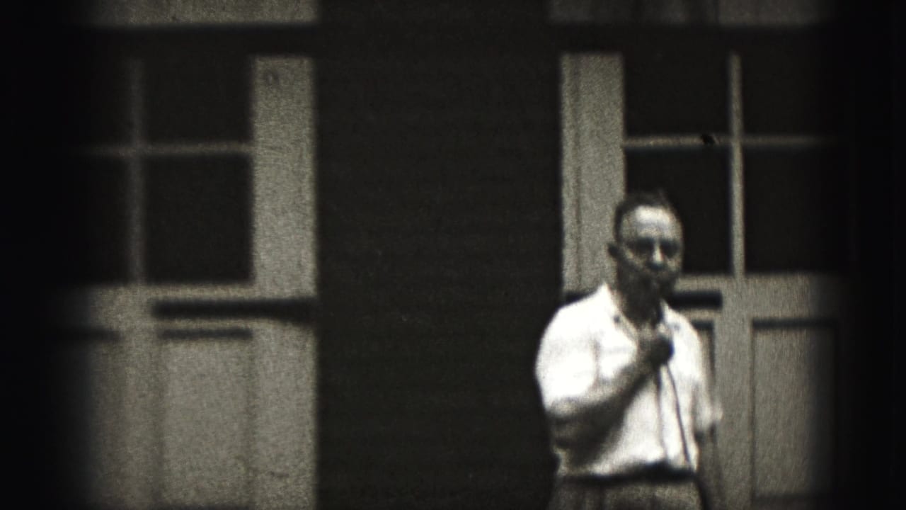 Man in glasses holds a microphone outside a building with multi-pane windows.