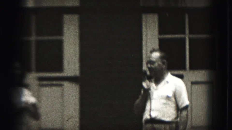 The man continues holding the microphone near the building while a blurred foreground figure partially obscures the 1946 New Cumberland view.