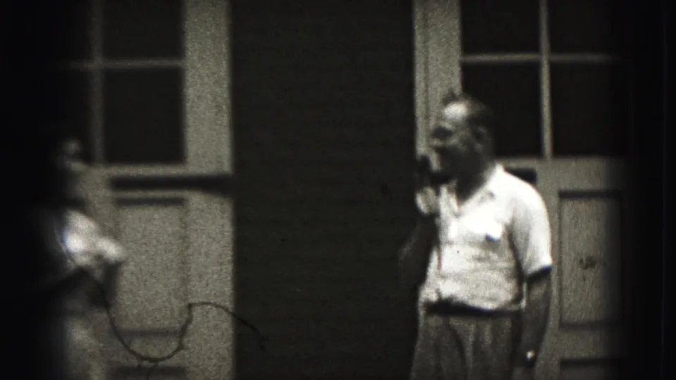A man in a light shirt holds a wired microphone outside the New Cumberland building, with cable and a partial foreground person visible.