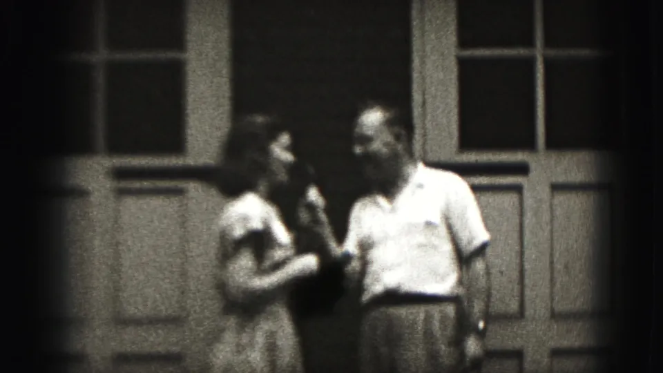 The man and woman stand near each other outside paneled doors, continuing a casual 1946 New Cumberland home-movie conversation.