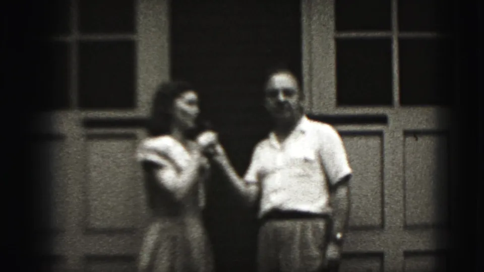 In 1946 New Cumberland home-movie footage, a woman in a light dress holds a microphone near a man outside paneled doors.