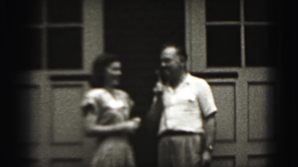 A man appears to speak into the microphone as the woman stands beside him outside the paneled doorway in 1946 home-movie footage.