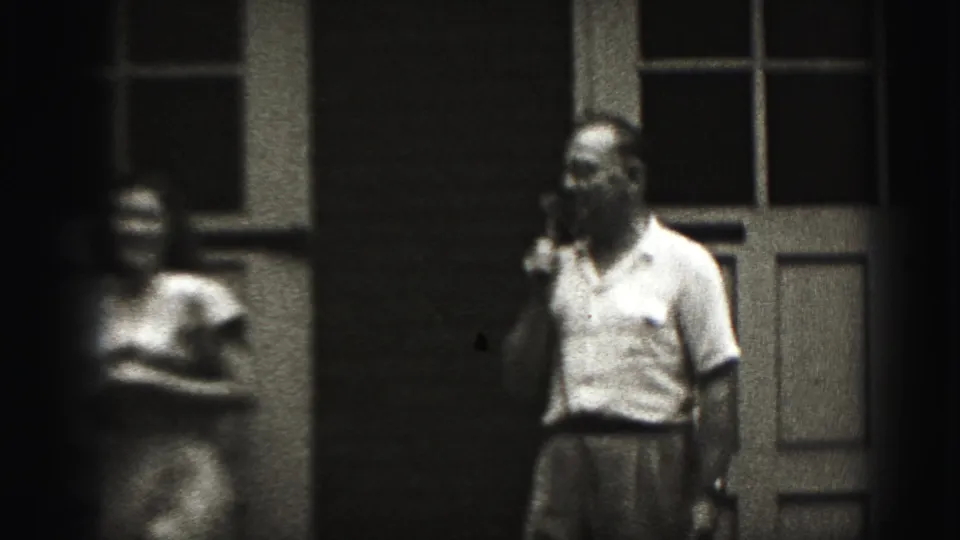 A man in a light shirt holds a telephone receiver outside while a woman stands to his left in soft-focus 8mm footage.