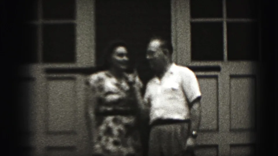 The couple remains outside the paneled doorway, with the woman turned toward the man in the 1946 home-movie scene.