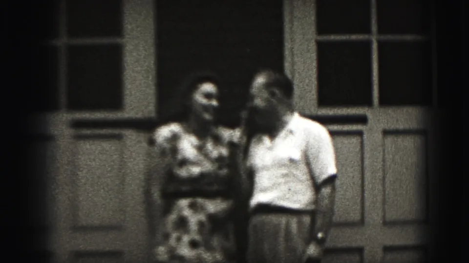 The couple remains close together outside the door and window, with the woman in a patterned dress and the man in a light shirt.