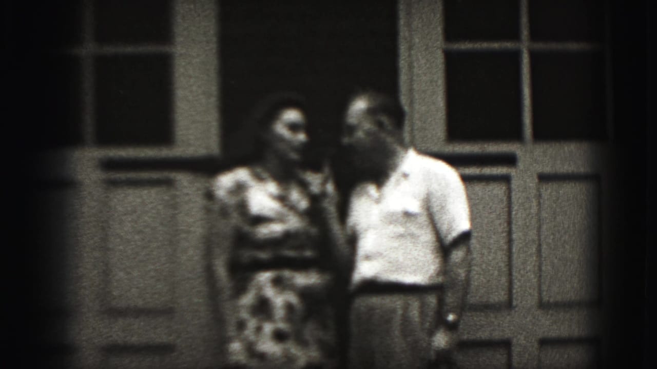 Adult couple standing close on a porch beside a window and paneled doorway.