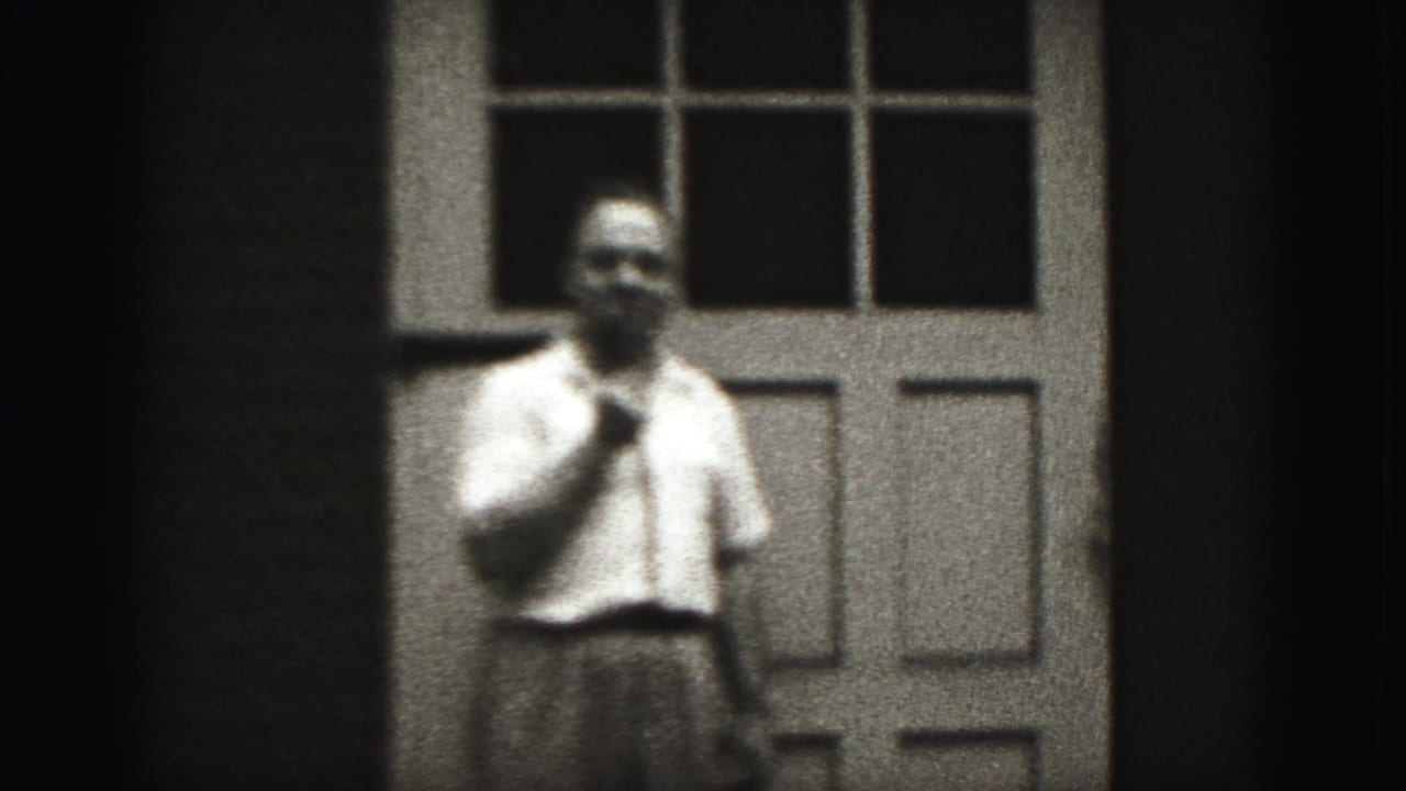 Adult man in 1946 New Cumberland home-movie footage stands by a paneled door, holding a small object near his mouth.