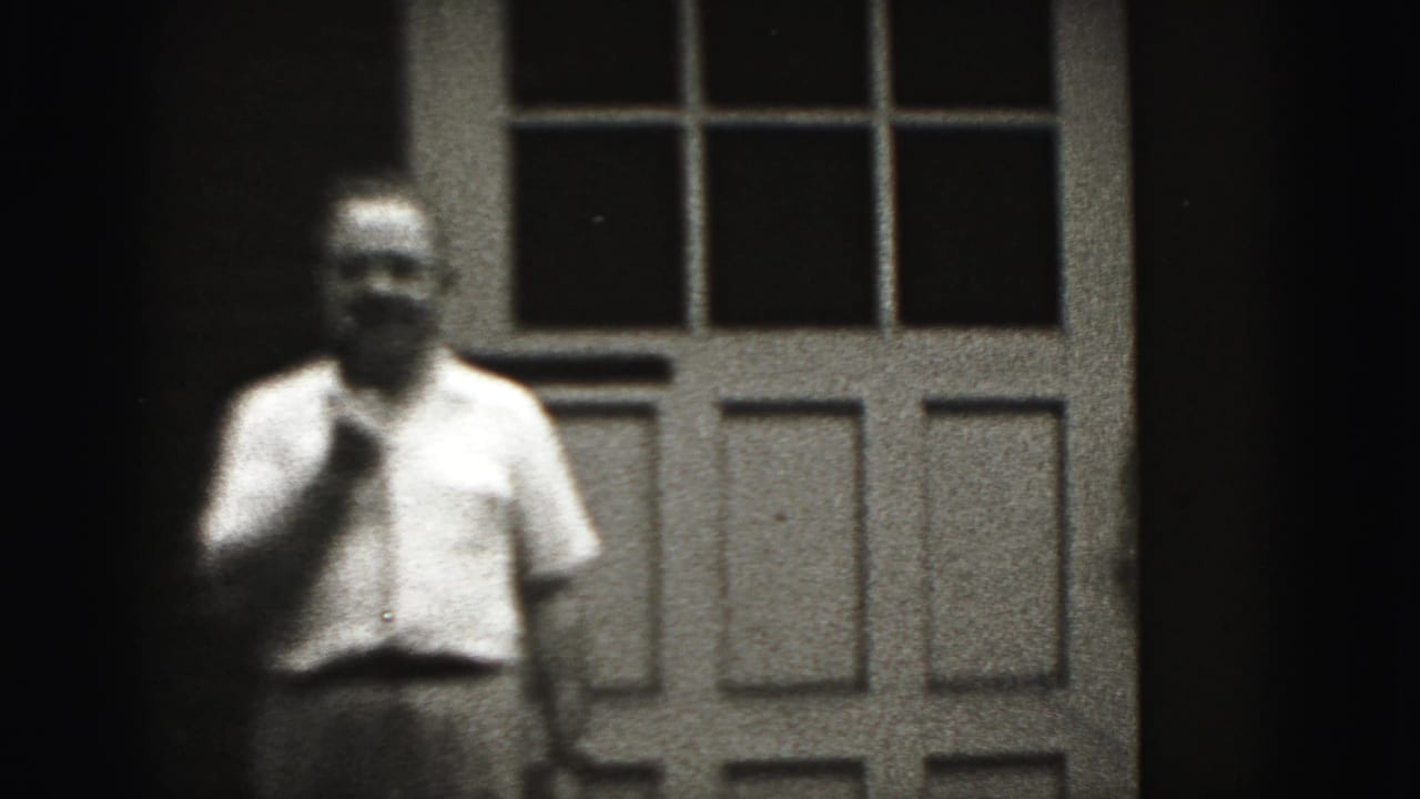 Adult man in a light shirt stands before the paneled doorway, softened by grain and focus in the 1946 home-movie frame.