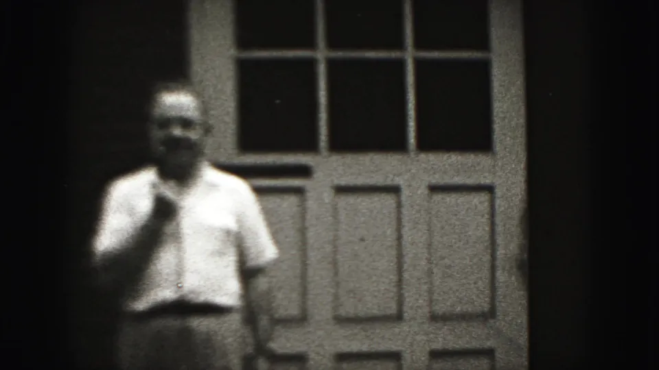 Adult man in a light shirt stands before the paneled doorway, softened by grain and focus in the 1946 home-movie frame.