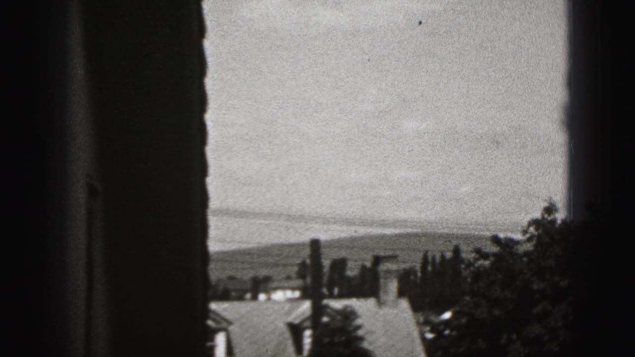 View through a window toward rooftops, trees, distant hills, and cloudy sky.