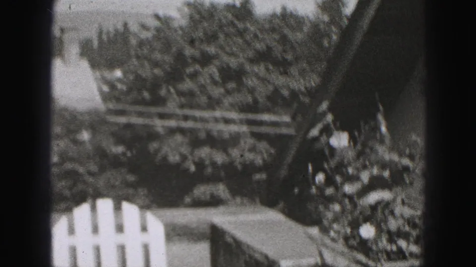Black-and-white 8mm frame appears to show a balcony or window view with white railing, trees, rooftops, and heavy edge shadow.