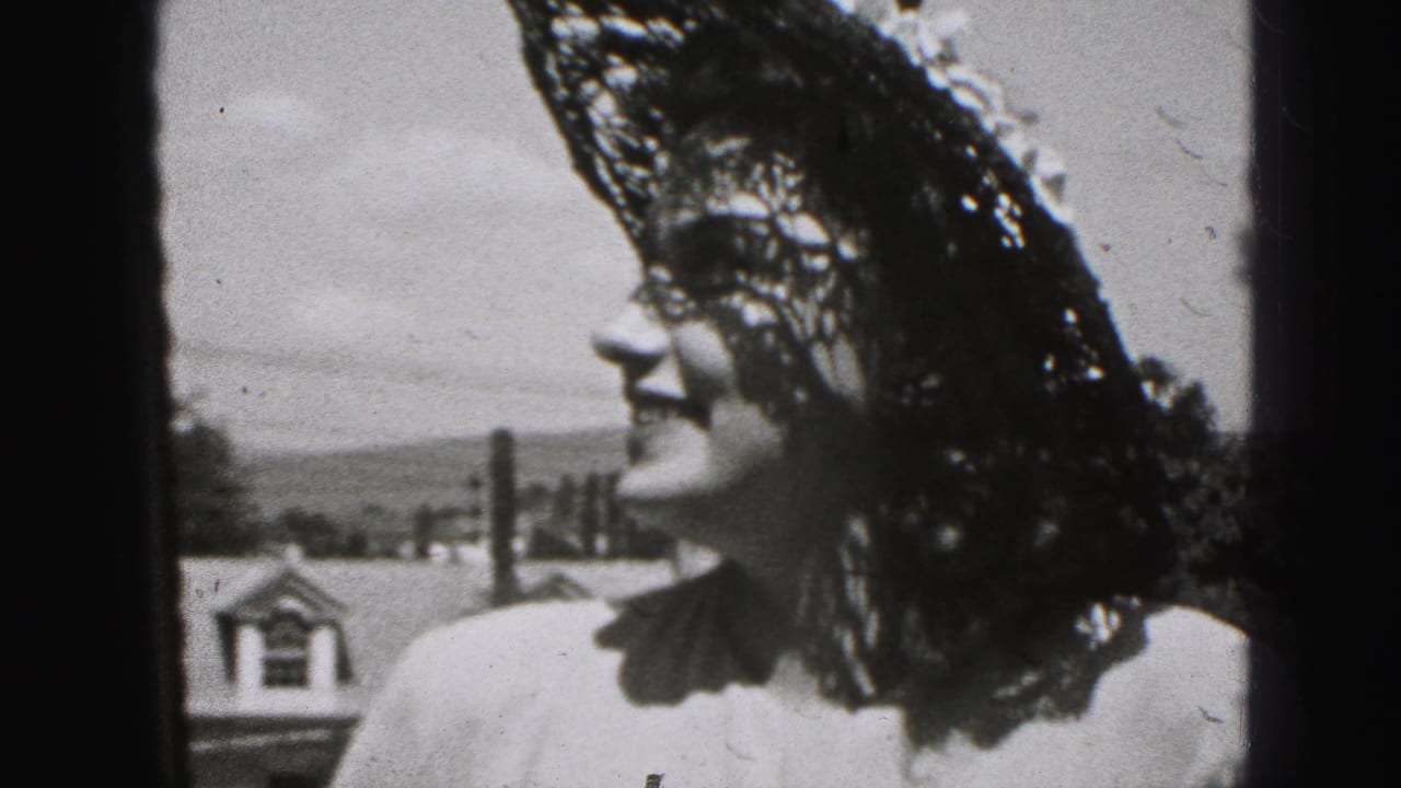 Black-and-white 8mm frame from New Cumberland shows an adult woman in a lace hat smiling in profile against rooftops and sky.