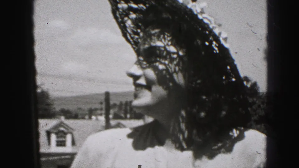 Black-and-white 8mm frame from New Cumberland shows an adult woman in a lace hat smiling in profile against rooftops and sky.