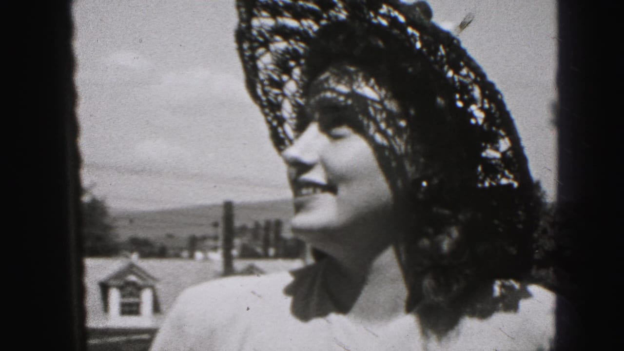 Woman in a lace hat smiles outdoors beside a roofline and distant hills.