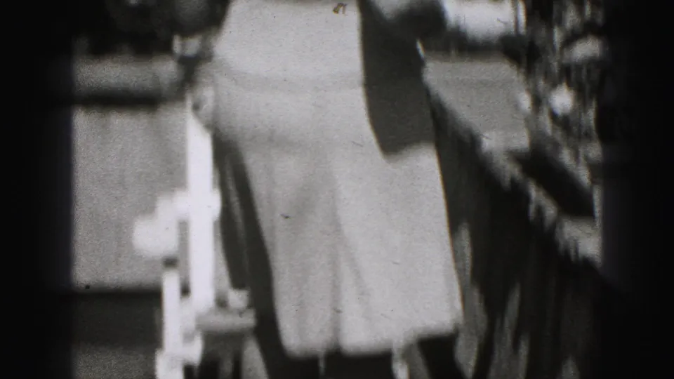 A partially framed woman in a light dress stands outdoors, with another figure cropped at the edge of the 1946 home-movie frame.