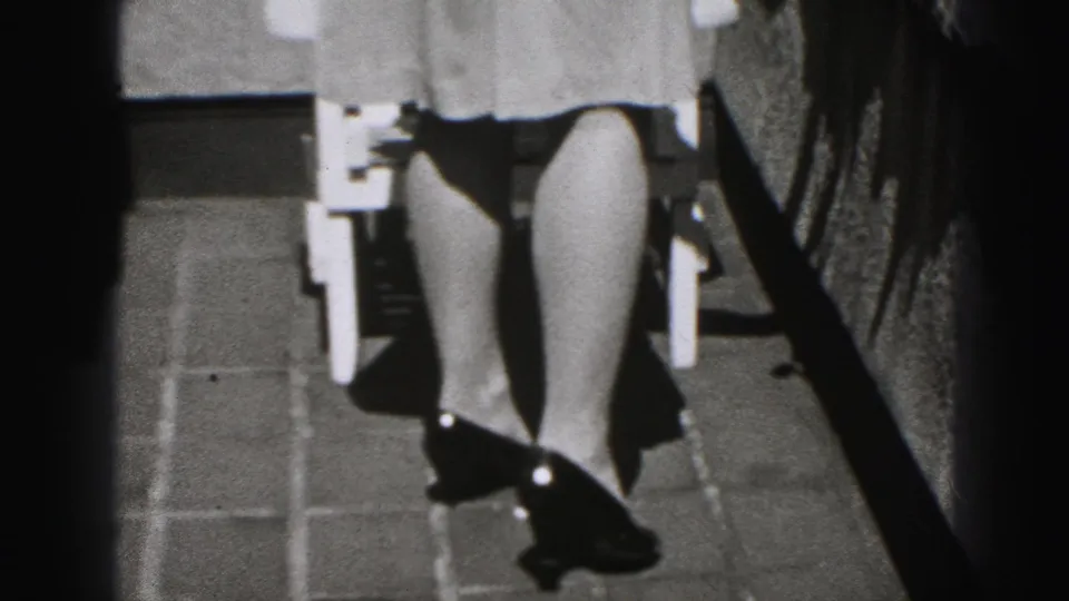 Black-and-white 1946 home-movie frame from New Cumberland showing a woman’s lower dress, legs, high heels, and part of an outdoor chair.