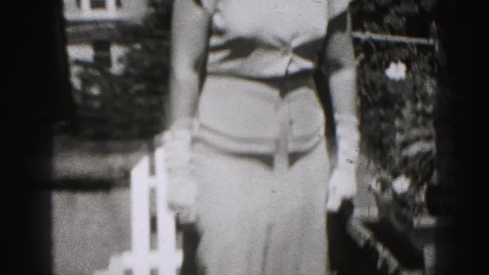Black-and-white 1946 frame showing a woman in a light dress and gloves posing outdoors near a white chair in New Cumberland.