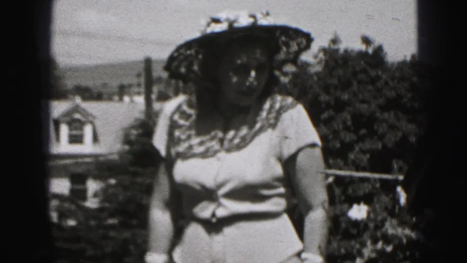 Black-and-white 1946 New Cumberland frame of a woman in a flowered hat, light dress, and gloves standing in a residential outdoor setting.
