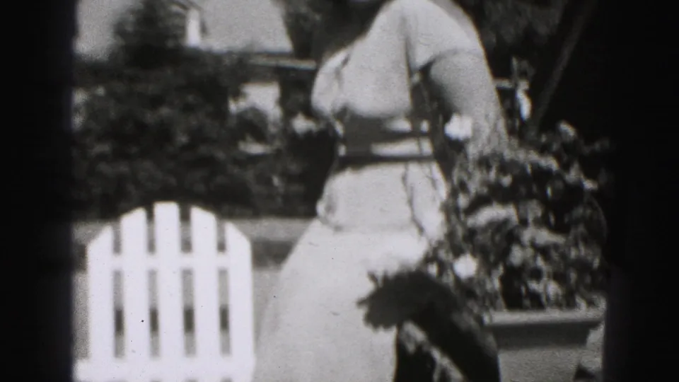 Black-and-white 1946 home-movie frame showing a woman in profile near shrubs and a white outdoor chair in New Cumberland.