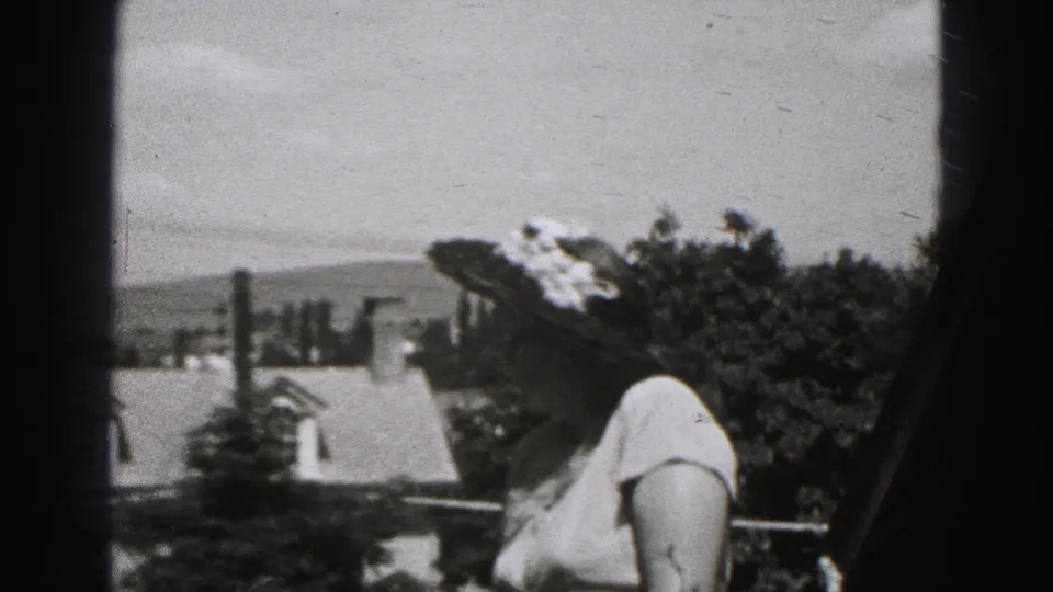Adult woman in a floral hat on a New Cumberland balcony, with houses, trees, and distant hills in view.
