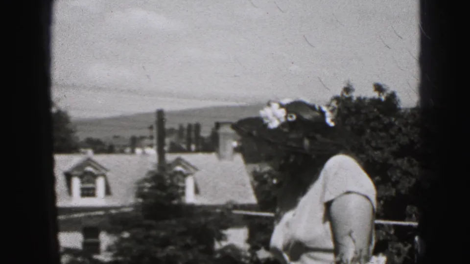 Side profile of the woman in a floral hat on a 1946 New Cumberland balcony, with residential roofs below.