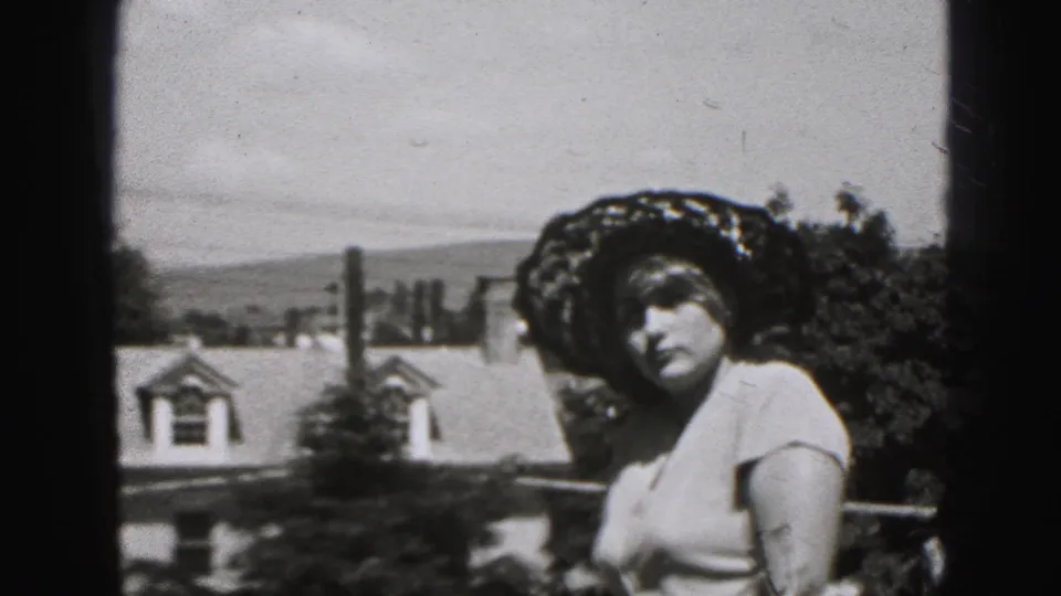 The woman turns toward the camera on a New Cumberland balcony, her floral hat and light blouse visible against rooftops.