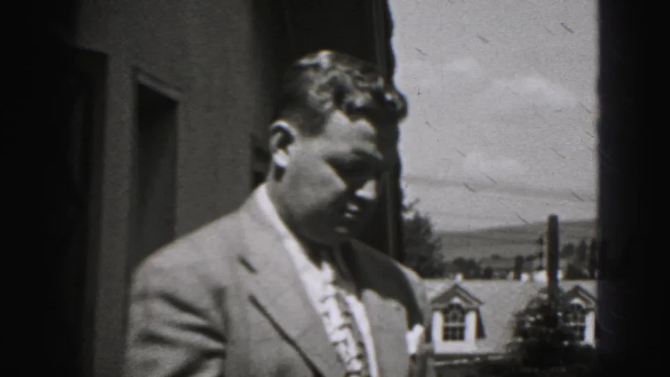 Man in a suit stands near a building in 1946 New Cumberland home-movie footage, with residential rooftops behind him.