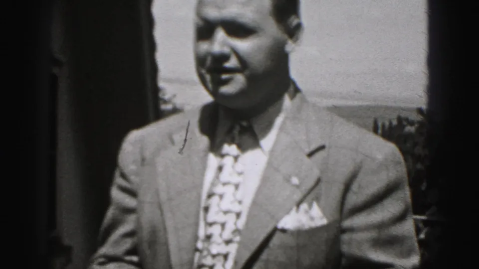 Close outdoor view of a suited man in 1946 New Cumberland home-movie footage, with a building edge and rooftops nearby.