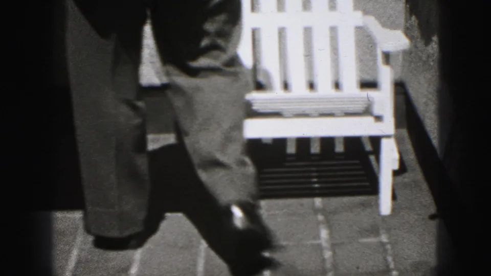 The man’s suit trousers and shoes are seen as he moves past the white chair along a paved outdoor path.