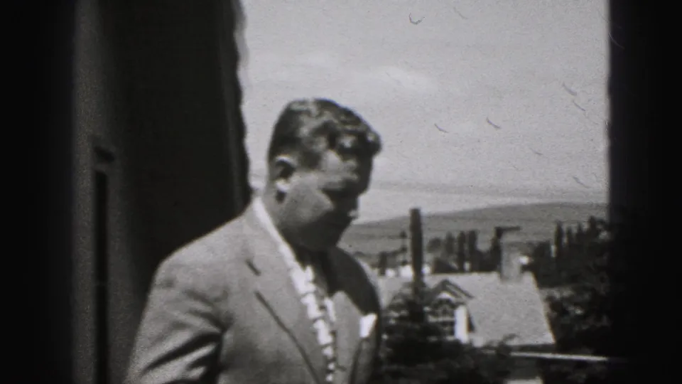 The man in a tailored suit turns sideways outdoors in New Cumberland, with a roofline, chimney, trees, and hills in the background.