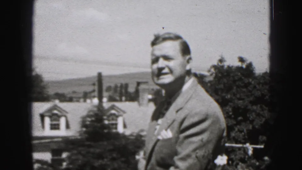 The man stands in partial profile outdoors, framed by a house dormer, chimney, trees, and distant hills in New Cumberland footage.