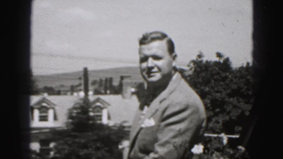 In 1946 New Cumberland, Pennsylvania footage, a suited adult man faces the camera outdoors near a railing, with rooftops and trees in the background.